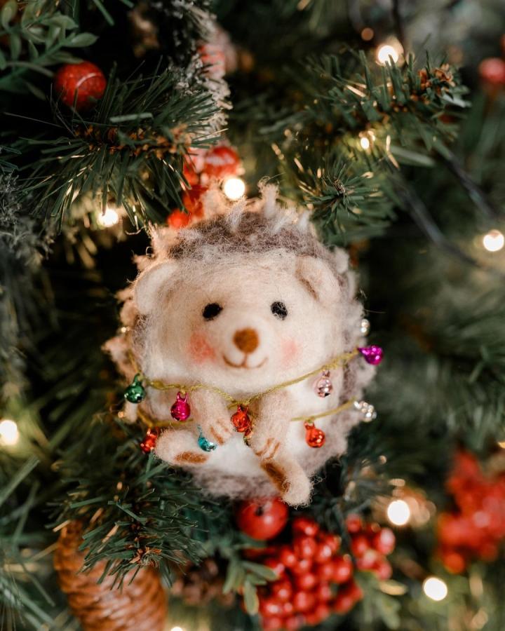 Cream and brown needle felted hedgehog ornament on Christmas tree with colorful tiny bells by @craftykitcompany. Soft wool texture with playful detail.