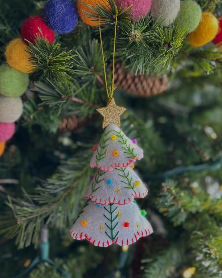 White felt layered Christmas tree ornament with colorful embroidery and beads hanging on a green pine branch handmade by @candidly.curious with cozy stitching details.