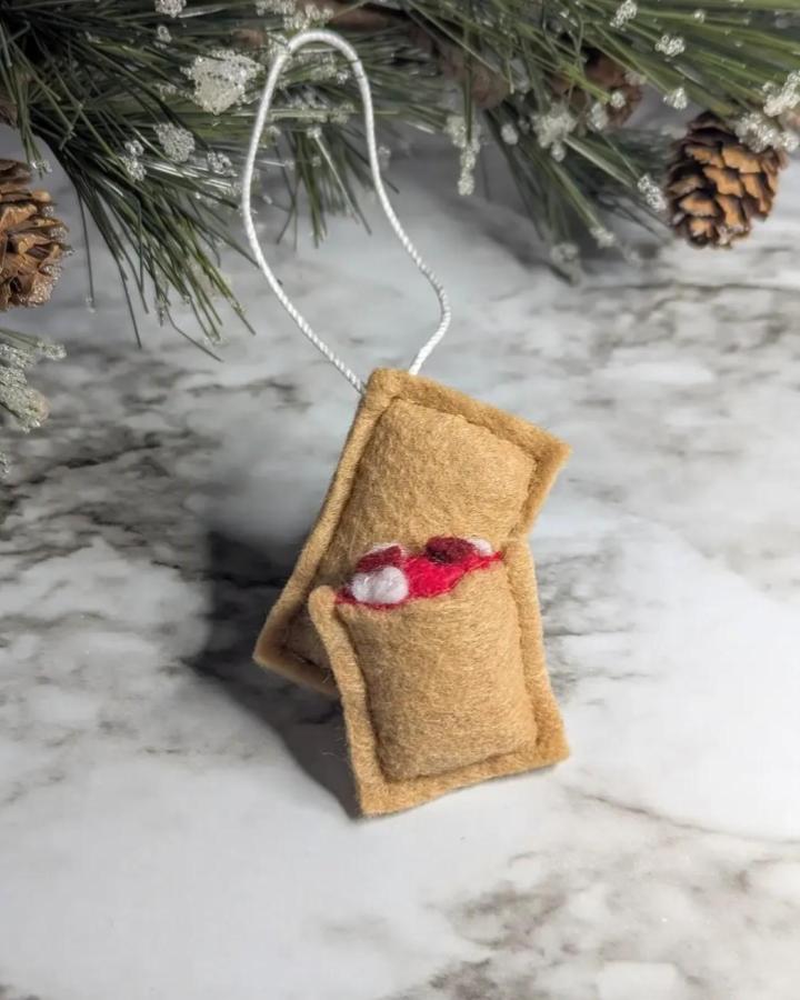 Tan felt pizza roll ornament inspired by cozy homemade crafts from @olivethisfelt on marble surface with pine and pinecones.