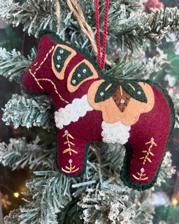 Burgundy and green felt handcrafted Scandinavian horse ornament from @the.christmaslady on a frosted pine branch with detailed embroidery.