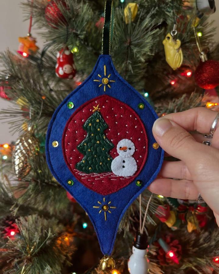 Blue and red hand-stitched felt ornament with snowman and Christmas tree from @meoandco.design hanging on Christmas tree. Festive textile craft detail.