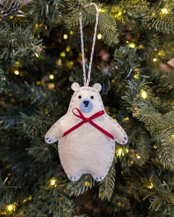 White felt polar bear ornament with hand-stitched details and red bow hanging on tree from @emilietrahan.illu, cozy handmade holiday decor.