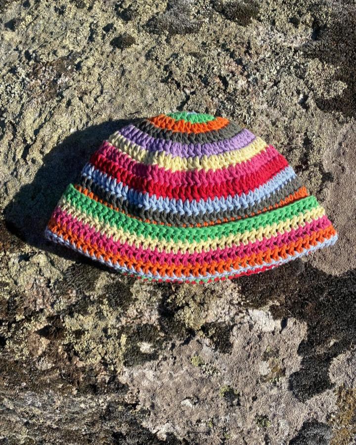 Colorful striped crochet bucket hat on textured rock surface from @tanaluarte. Bright yarn and bold playful stitching design.