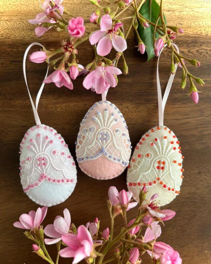 Soft pastel felt lace Easter egg ornaments decorated with beads from @nata_d_craft on wooden surface with pink flowers. Charming handmade spring decorations.