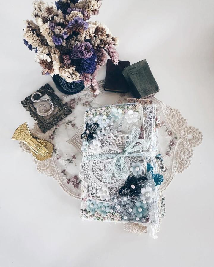 White and pastel blue lace junk journal with floral embellishments on vintage lace doily by @berrymixjournals. Delicate fabric and bead details enhance the handmade aesthetic.