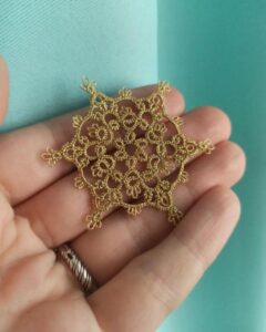 Golden miniature tatting lace piece held in hand from @bardar_svetlana_lace. Delicate loops and picots forming a star pattern.