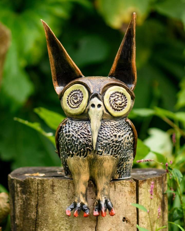 Brown and green textured owl sculpture with exaggerated features on tree stump by @livefirecook. Handcrafted with detailed feather patterns and whimsical eyes.