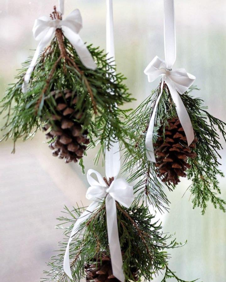 Brown pine cones with green cedar and pine sprigs tied with white satin bows hanging from ribbons by @marie.levang. Natural rustic indoor decor idea.