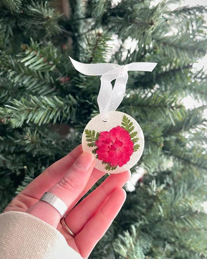 Pink flower and green fern resin ornament held near Christmas tree from @themorningglowstudio. Handmade holiday decoration with clear shimmering resin.