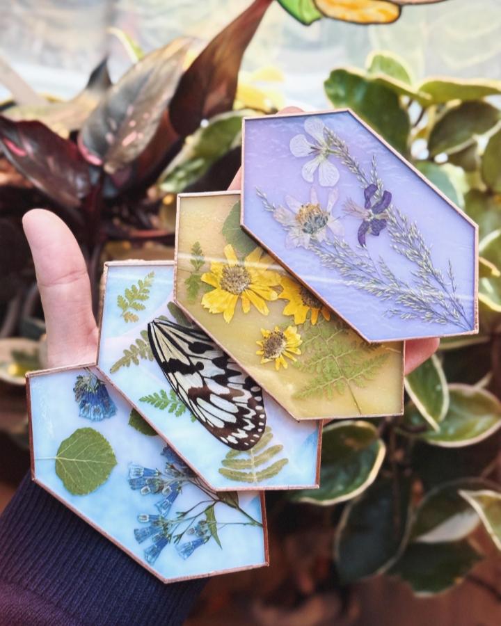 Colorful stained glass-inspired pressed flower art pieces by @jessbreaksglass featuring detailed floral and fern arrangements in geometric copper frames.
