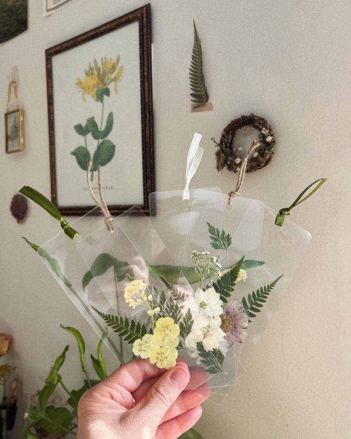 Yellow white and purple pressed flower bookmarks laminated with fern details from @wingandroot held against a neutral wall background. Simple handmade floral craft accessories.