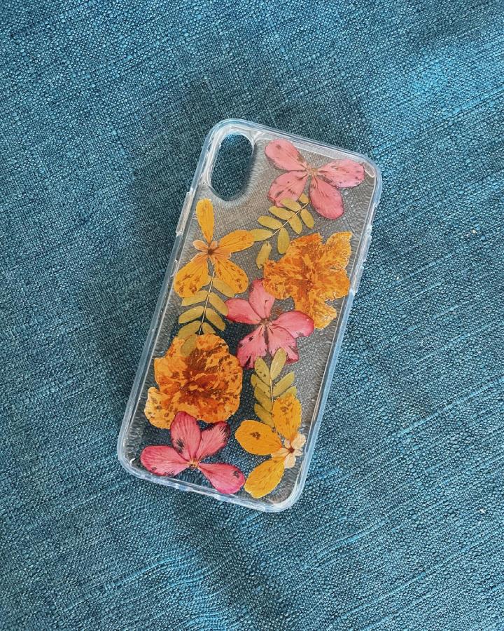 Clear phone case decorated with orange and pink pressed flowers and leaves from @flowerbandit.design displaying creative pressed flower art ideas.