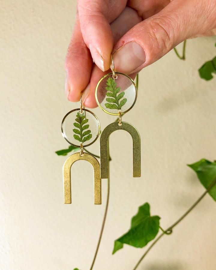 Green fern encased in clear resin with gold metal arches handmade earrings from @theretrocollectiveco showing nature-inspired jewelry craft.
