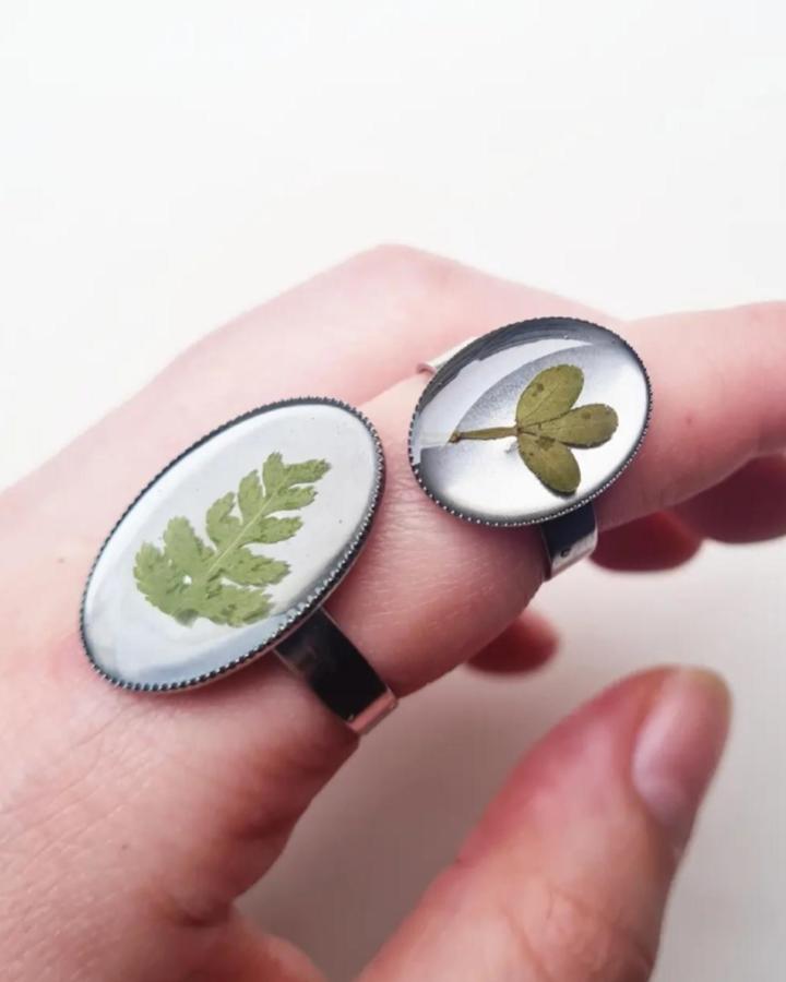 Green pressed leaves encased in resin rings by @vicky.createsstuff. Close-up of delicate botanical jewelry on fingers.