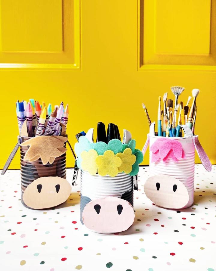 Playful cow-inspired tin can crafts from @miss_yellow_shoes with felt and paint details holding art supplies. Bright yellow background.