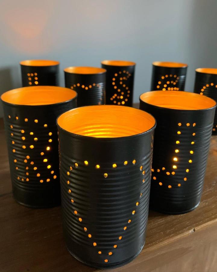 Black and orange painted lantern cans by @homecycleduk with dotted letter designs glowing softly on a wooden surface.