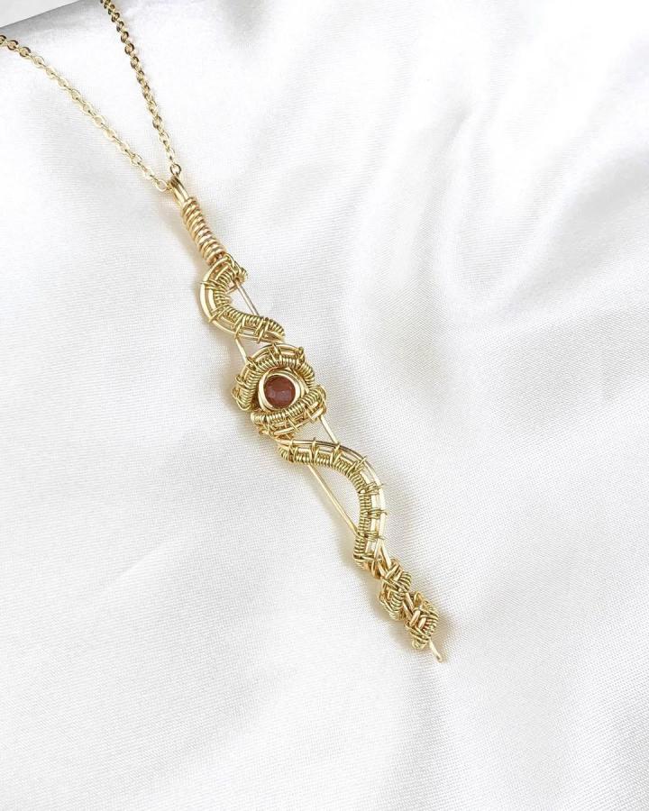 Gold wire wrapped garnet dagger pendant necklace from @rediscoveryourlightjewelry on white fabric showcasing detailed handcrafted metalwork jewelry.