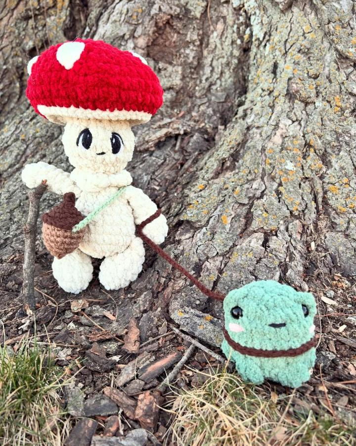 Red and cream plush mushroom crochet character with mint-green pet and acorn accessory by @cluckandbuzzcrochet. Forest-inspired playful amigurumi design.