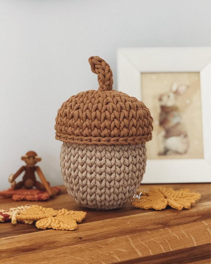 Chunky brown and beige crochet acorn with twisted stem from @merinocatcreation on wooden surface with autumn leaves and decor.