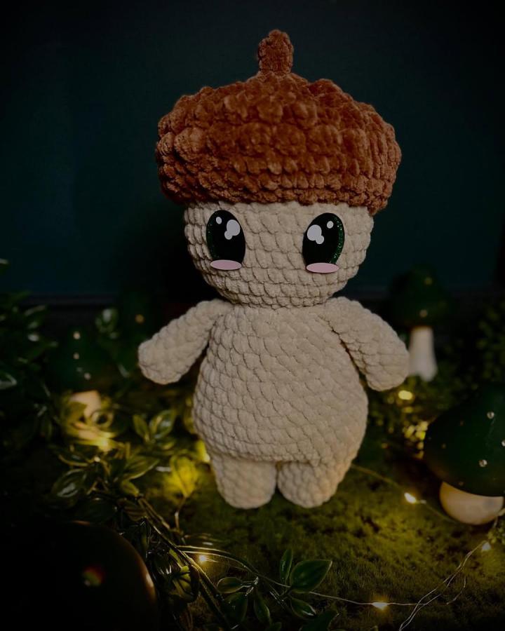 Soft plush beige and chestnut acorn crochet doll by @hanner_gurumi with textured stitch and cute large eyes surrounded by fairy lights and greenery.
