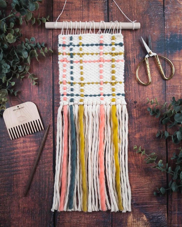 Cream yarn plaid wall hanging with coral, mustard, and charcoal stripes by @spruceandlinen showcasing unique yarn wall hanging ideas. Chunky texture and long fringes.