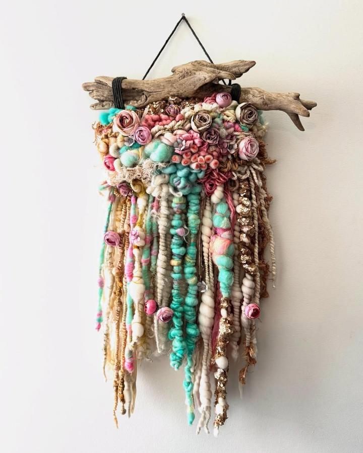 Soft pastel fiber art wall hanging with driftwood base from @mothmetal. Features chunky yarn, sequins, and floral accents.