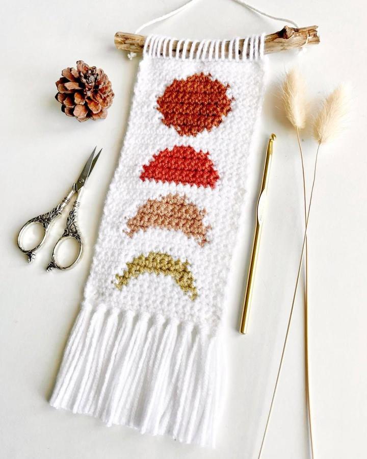 White crochet wall hanging with burnt orange and green shapes on driftwood by @loveandstitchh. Cozy handmade textured decor with fringe.