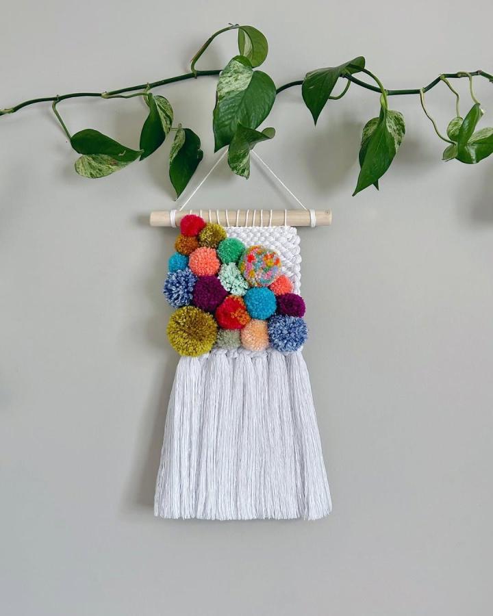 Colorful pom pom and fringe wall hanging from @therobinsnest_. Bright yarn balls on white woven base with wooden dowel hanger.