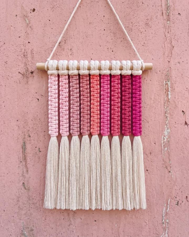 Pink and peach gradient cotton yarn tassel wall hanging from @macrameforbeginners with wooden dowel on textured pink wall.