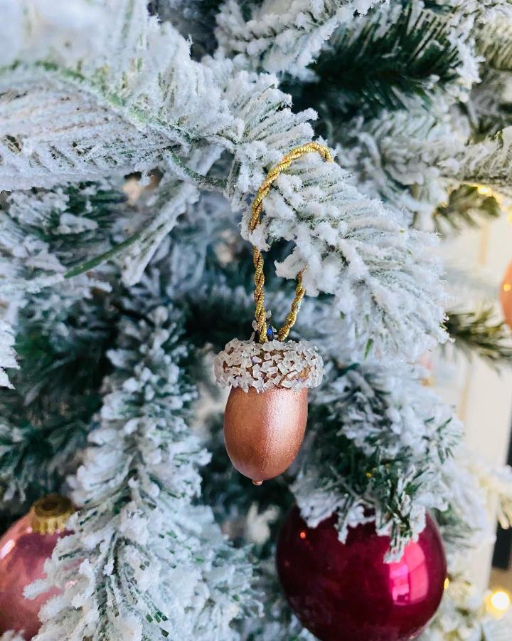Rose gold glittery acorn ornament with white crystals hanging on snowy evergreen branch from @sohabeachglass. Holiday craft with natural elegance.