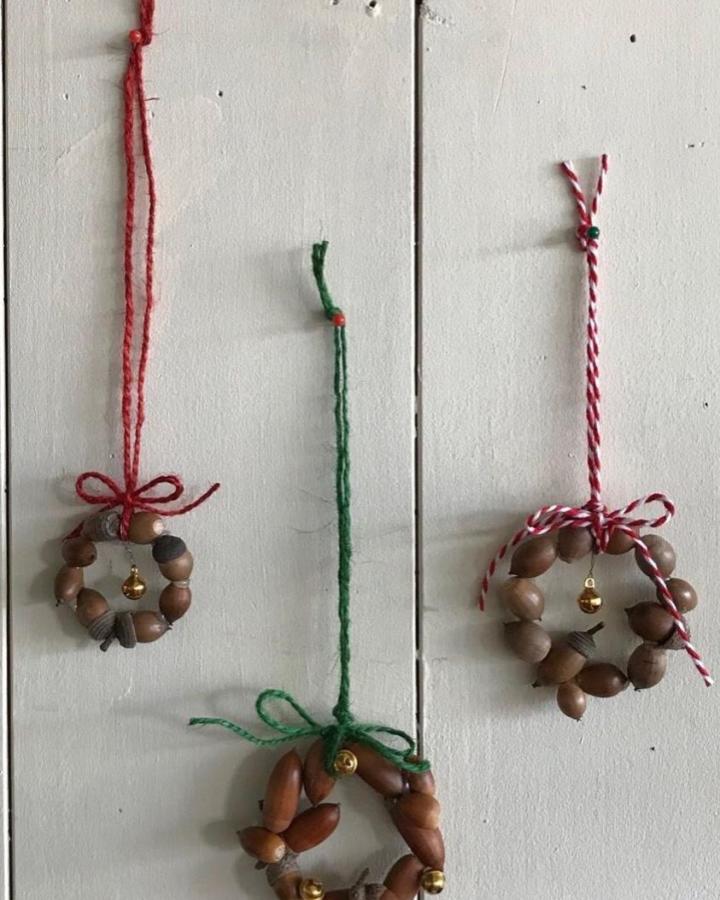 Natural acorn wreath ornaments with colorful twine and tiny gold bells from @gomatokyo hanging on white wood background. Simple festive craft idea.