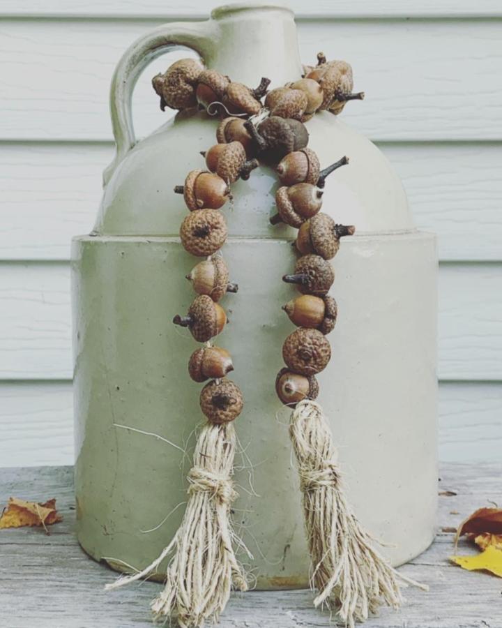 Rustic brown acorn garland with natural twine tassels displayed on a vintage jug from @wisconsinmagpie. Natural autumn-inspired craft decoration.