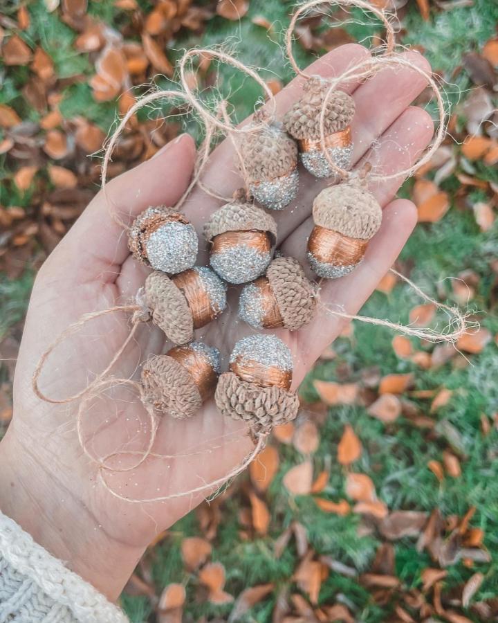 Natural acorn ornaments dipped in copper wire with silver glitter detail by @raising.little.flowers shown on a hand outdoors. Rustic seasonal craft idea.