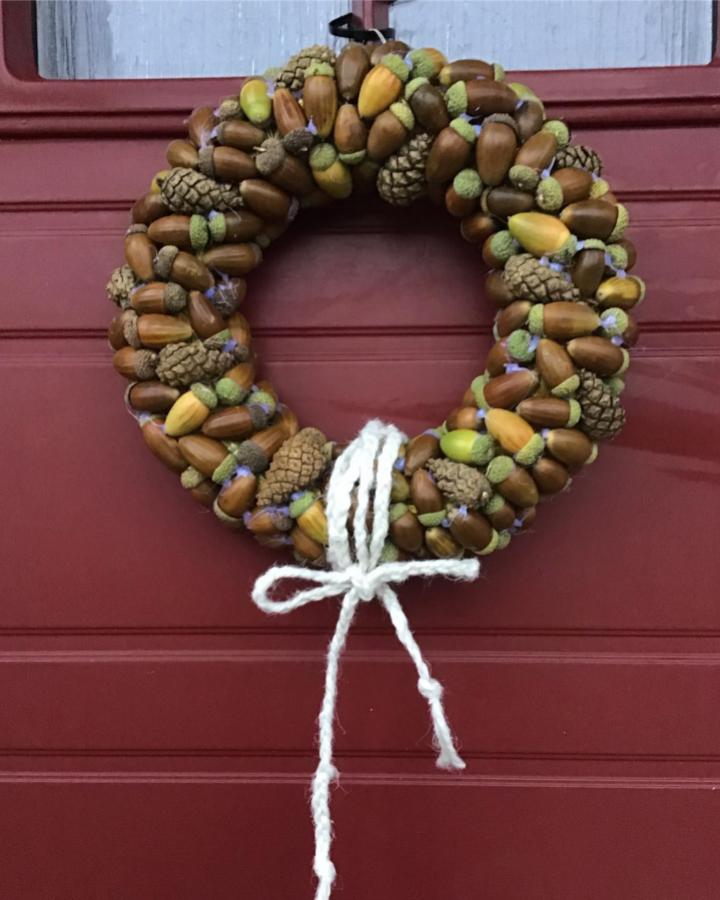 Brown and green acorn wreath with pinecones and white yarn bow on red door by @svartenkatt003 natural autumn decor craft.