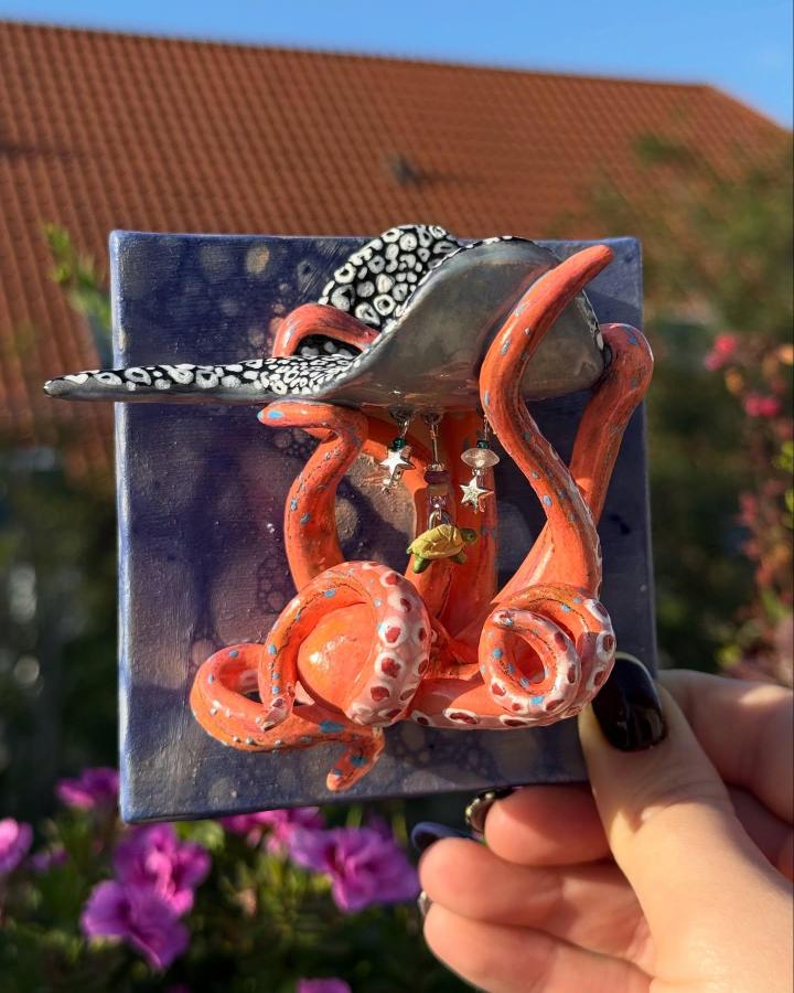 Bright coral orange octopus sculpture with dangling charms on deep blue hand-painted tile by @studiomasomaso. Whimsical marine-themed ceramic art.