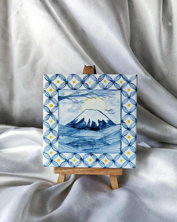 Blue and yellow hand-painted decorative tile on wooden easel by @riaastudio featuring a mountain landscape and geometric border pattern.