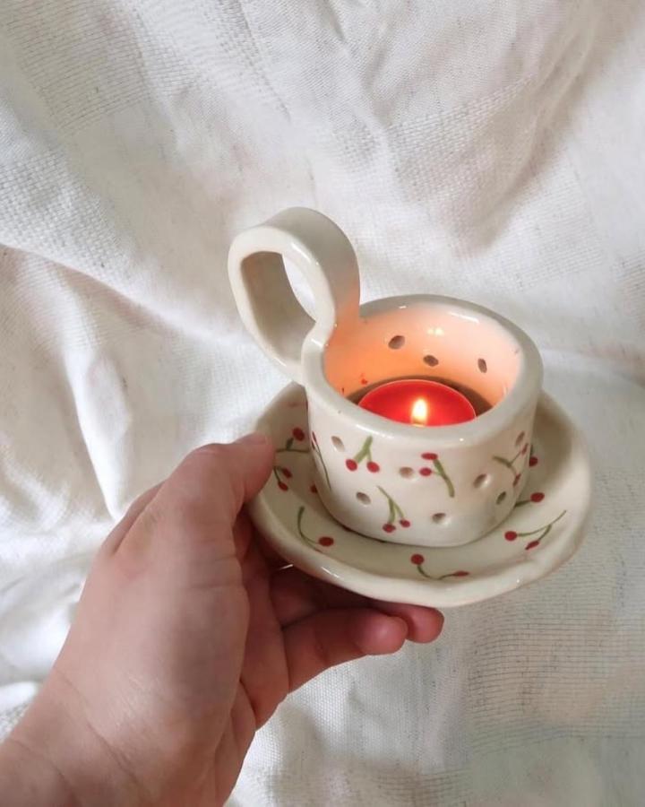 Creamy white ceramic teacup candle holder with red cherry details and perforated light holes by @coral.ceramics. Handcrafted cozy charm accessory.