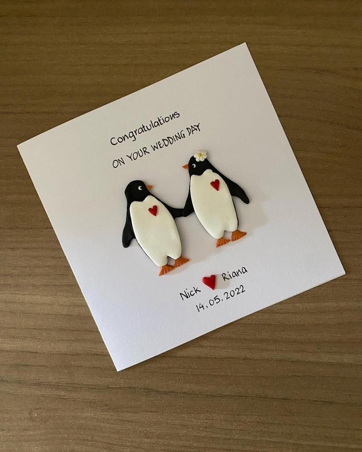 Black and white polymer clay penguin couple on a white card from @bakemeacard. Cute handcrafted wedding greeting with tiny red hearts and flower.