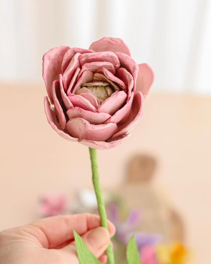 Pink handcrafted egg carton flower held by fingers showcasing layered petals and green stem for DIY home decoration.