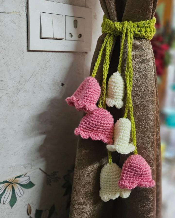 Pink and cream crochet flower curtain ties with green yarn from @crochet_by_priya_ hanging on brown curtain adding handmade floral charm.