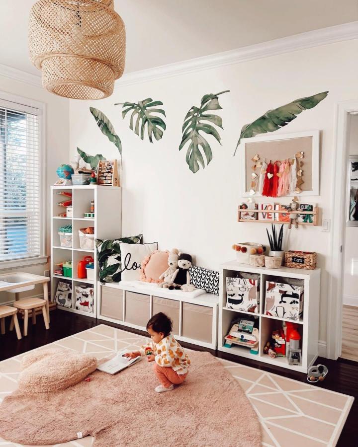 White modular Kallax bench with soft cushions and storage bins in a bright kids playroom by @thismamaloveslife. Doubled shelving units and playful leaf wall decals create cozy organized space.