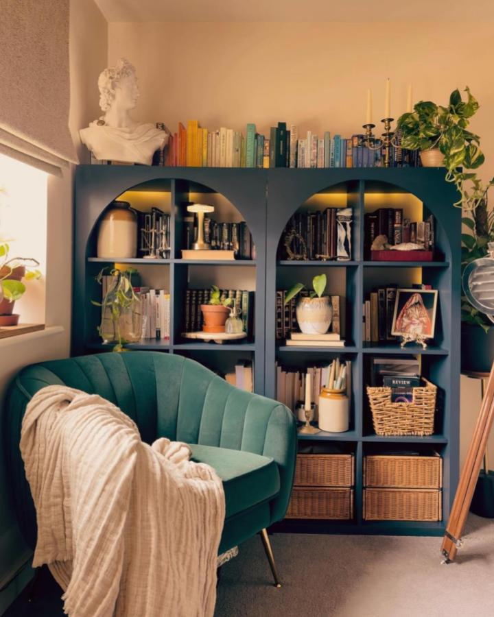 Dark teal arched shelving unit with warm lighting and baskets from @myrevampreveal Cozy home library display with plants and books.