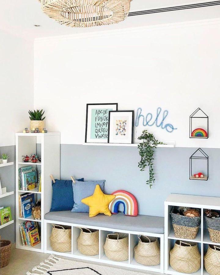 Natural white shelving unit with gray cushioned bench and wicker basket storage for kids room from @playroominspo. Cozy, colorful, and organized play area.