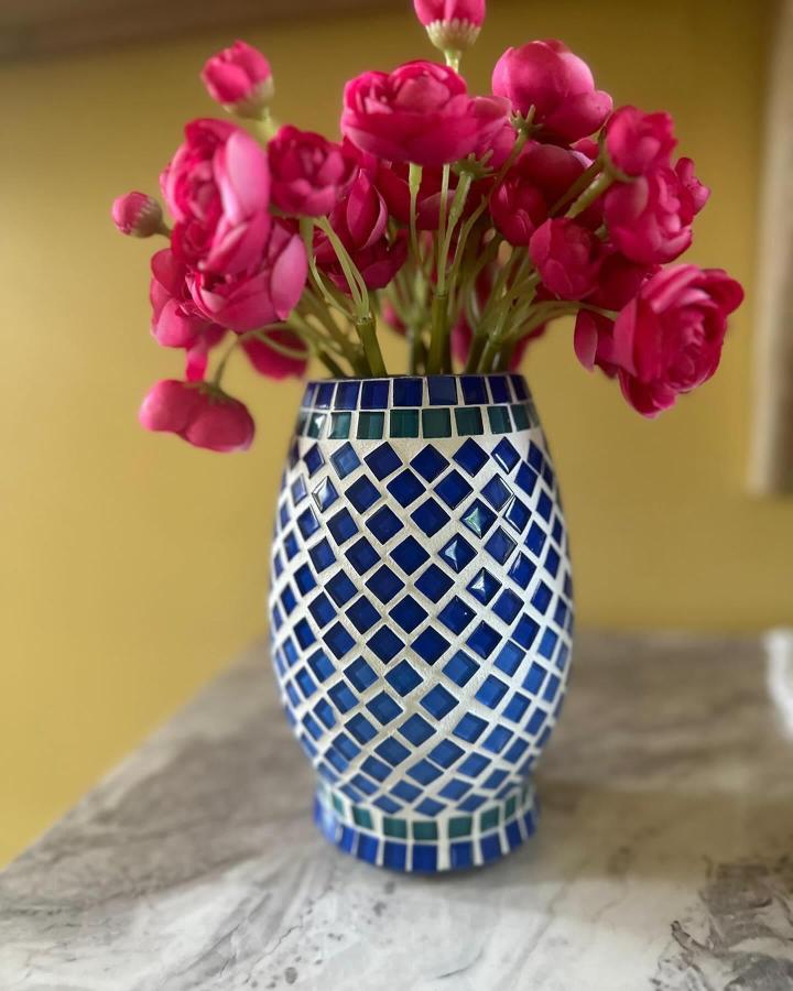 Blue and teal glass tile mosaic vase from @mirrorsmosaicmagic with diamond pattern and bright pink flowers on marble surface.