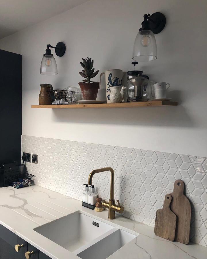 Warm wood floating shelf above white geometric backsplash and brass faucet from @lyttelton_interiors. Rustic kitchen storage with plants and ceramics.