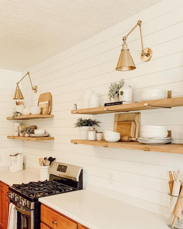 Warm wood floating shelves with brass lighting in a bright kitchen by @ambereveryday_ displaying white ceramics and greenery in a cozy rustic style