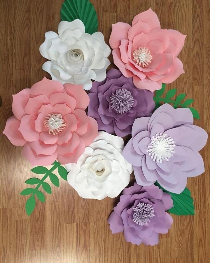 Pastel layered paper flowers with green leaves on wooden floor from @aprendefloresdepapel. Large, detailed handmade blooms.
