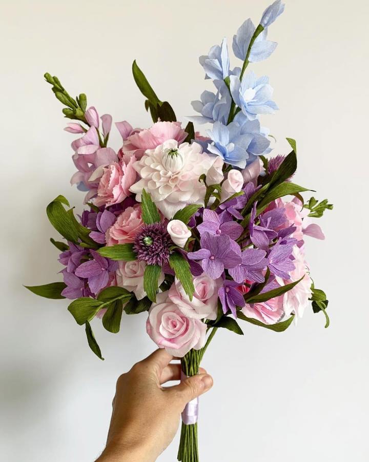 Pastel crepe paper floral bouquet held by hand from @petalandbird. Soft pink, purple, and blue blossoms with green leaves in delicate arrangement.
