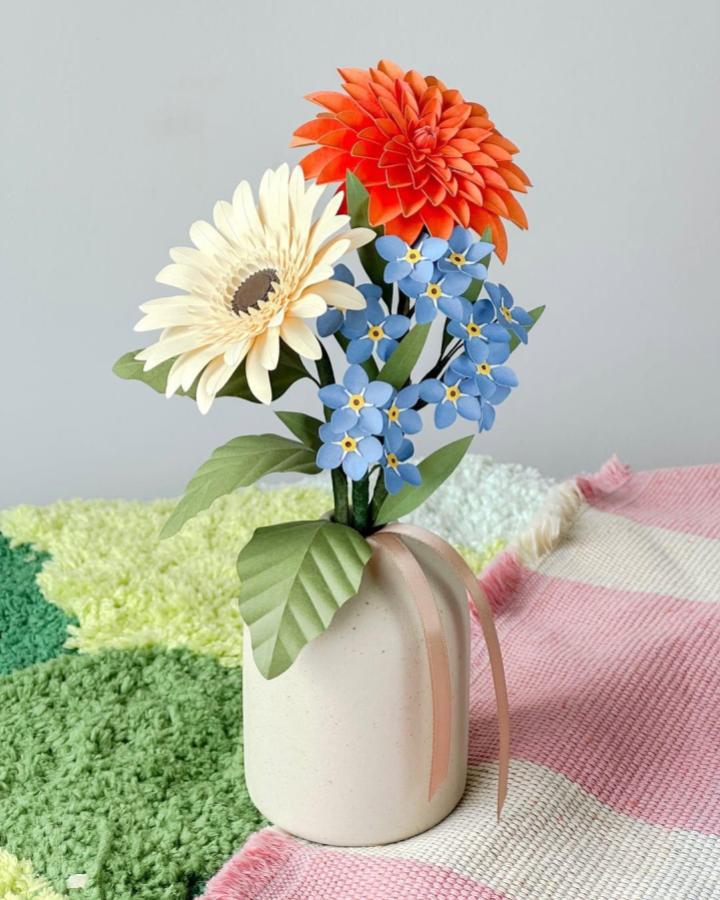 Orange dahlia, creamy daisy, and blue forget-me-not paper flower crafts for adults in white vase from @paperbloom.studio. Bright and detailed handcrafted bouquet.