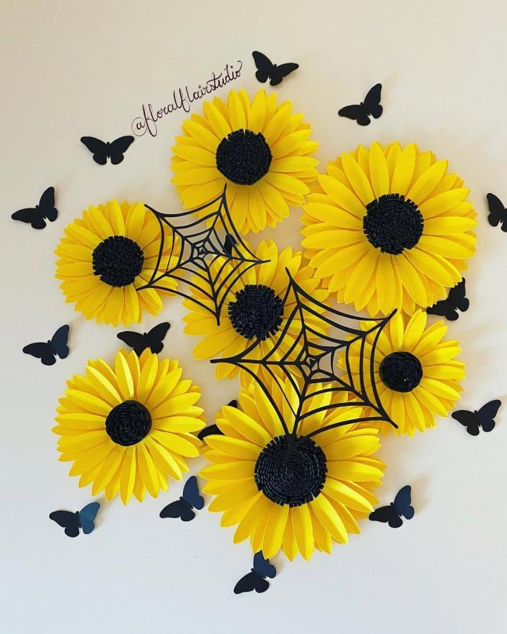 Bright yellow and black paper sunflowers with spider web and butterfly accents by @floralflairstudio. Vibrant floral wall art with spooky charm.